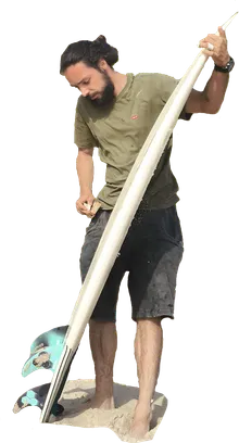 Hugo waxing a surfboard on the sand, board propped nose-up against his shoulder